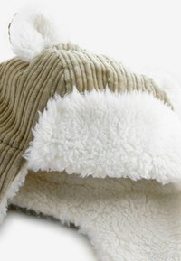 Tan corduroy-hattu, jossa on valkoista fleece-vuoria ja korvaläppät, joissa on pehmeät, pyöreät korvakoristeet ja teksturoitu, ribattu muotoilu.