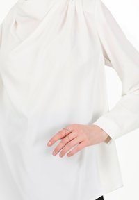 Hvid skjorte med draperet design, der har lange ærmer og en høj halsudskæring. En hånd prydet med delikate guld ringe hviler mod stoffet.