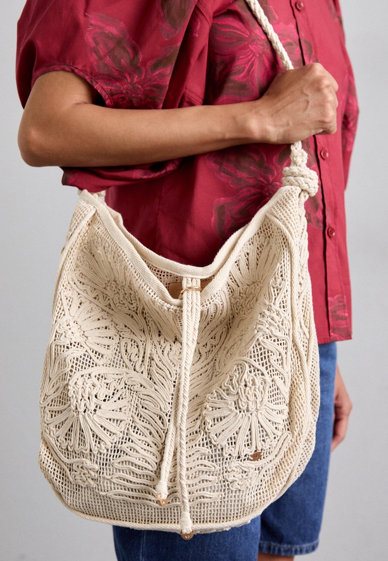 Macramé-Handtasche in Creme mit kunstvollen floralem Muster, Zugbandverschluss und geflochtenem Griff, kombiniert mit einem roten Blumenshirt.