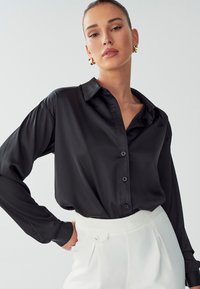Blusa in satin nero con colletto classico, maniche lunghe e chiusura a bottoni. Abbinata a pantaloni bianchi a vita alta. Texture liscia.