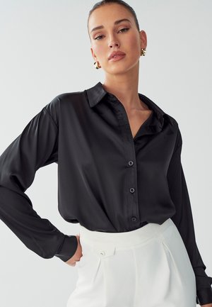 Black satin blouse with a classic collar, long sleeves, and button closure. Paired with high-waisted white trousers. Smooth texture.