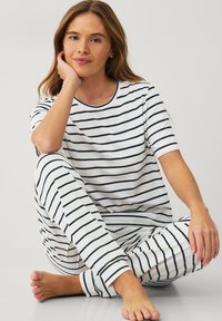 Kvinde med langt hår iført hvide pyjamas med sorte vandrette striber, siddende barfodet på gulvet og hvilende hovedet på sin hånd.