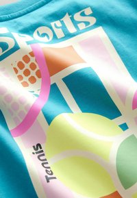 Turkise bomulds T-shirt med et grafisk design, der har sportsmotiver, herunder tennisbolde og geometriske mønstre i pink, gul og orange.