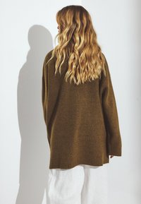 Suéter verde oliva de gran tamaño con puños y dobladillo acanalados, textura suave, corte holgado, usado sobre pantalones claros.