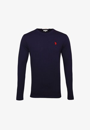 Mann trägt ein marineblaues Langarmshirt mit einem roten Logo, blaue Jeans und schwarze Turnschuhe mit weißen Schnürsenkeln, steht vor einem einfarbigen Hintergrund.
