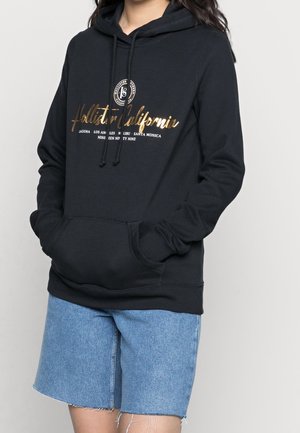 Persoon in een zwarte Hollister California hoodie met gouden en witte tekst, gecombineerd met lichtblauwe denim shorts met rafelige randen.