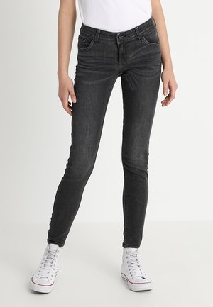 Kobieta w obcisłych, ciemnoszarych dżinsach i białych trampkach za kostkę, stojąca na neutralnym, jasnym tle.