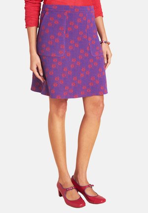 Frau trägt einen knielangen lila Rock mit roten Blumenmustern, ein rotes Langarmshirt, passende rote Pumps und ein Perlenarmband.