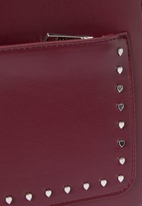 Sac à main en cuir bordeaux avec une poche avant, agrémenté de clous en forme de cœur argentés le long du bord et d'un fermoir en métal élégant.