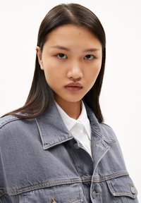 Chaqueta de mezclilla gris con cuello clásico, doble bolsillo frontal y costuras visibles. Lleva una camisa blanca de botones debajo. Diseño sencillo.