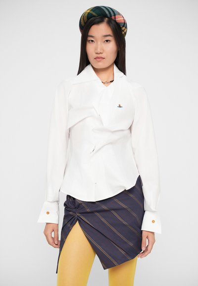 Witte, gekreukelde overhemdbloes met lange mouwen, geborduurd logo, gecombineerd met een gestreepte asymmetrische rok en gele maillots.