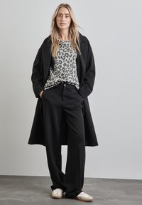 Cappotto lungo nero, camicia grigia con stampa leopardata, pantaloni neri su misura e sneakers bianche. Sono visibili texture lisce e pattern a contrasto.