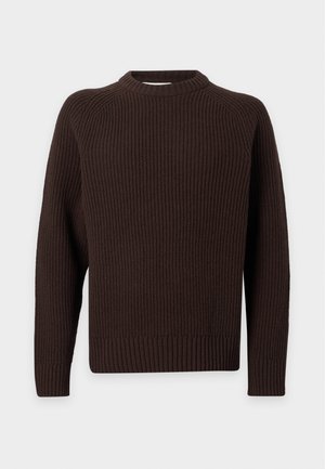 Homme portant un pull en maille marron foncé, un jean bleu clair ample et des chaussures marron foncé, debout avec une main dans la poche.