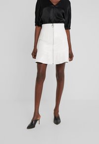 Femme portant une mini-jupe en cuir blanc taille haute avec fermeture éclair à l'avant, un chemisier en satin noir, et des mules à talons noires à bout pointu.