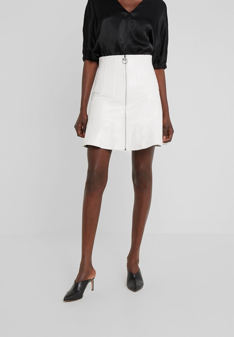 Femme portant une mini-jupe en cuir blanc taille haute avec fermeture éclair à l'avant, un chemisier en satin noir, et des mules à talons noires à bout pointu.