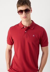 Rotes Poloshirt mit Kragen und kurzen Ärmeln, mit einem kleinen blauen Logo auf der Brust. Modell trägt dunkle Sonnenbrillen.