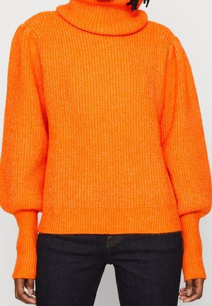 Personne portant un pull à col roulé en maille épaisse orange vif avec des manches ballon et un pantalon foncé sur fond neutre.