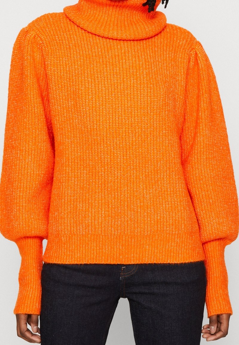 Personne portant un pull à col roulé en maille épaisse orange vif avec des manches ballon et un pantalon foncé sur fond neutre.