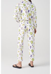 Conjunto de loungewear blanco con patrones divertidos de caras sonrientes en verde, amarillo, púrpura y rosa. Presenta una chaqueta corta y pantalones de corte relajado.