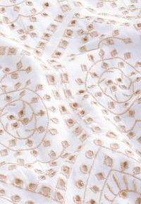 Hvidt stof med beige broderede blomster- og geometriske mønstre, der har tekstureret syning og udskårne detaljer i en flydende sammensætning.