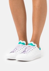 Weiße Sneakers mit einer glatten Lederoberfläche, lila Schnürsenkeln und türkisfarbenen, gewellten Akzenten an der Ferse. Mit perforierten Details an den Seiten.