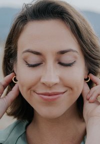 Cercei hoop de aur purtați de o persoană cu păr scurt, ondulat și castaniu. Fundalul arată un peisaj natural neclar.