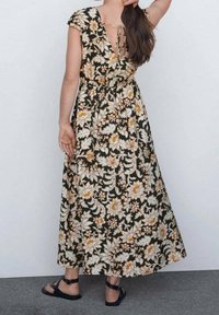 Maxikleid mit einem dunklen Blumenmuster in Creme und Orange. Es verfügt über kurze Kappenärmel, eine Schnürung am Rücken und eine fließende Silhouette.