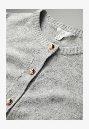 Heled hall kardigan pehmest kangast. Omab ribadeed kaelust ja kuldse tooniga nööpe esiosas. Sildil on nähtav kaeluses.