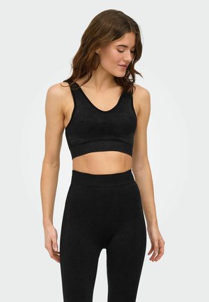 Kvinne iført svart ermeløs crop top og høytlivsleggings, stående og ser ned mot enkelt bakgrunn.