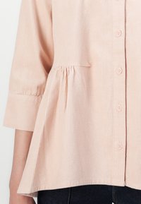 Blusa in corduroy rosa chiaro con design a peplum, maniche a tre quarti e cinque bottoni lungo il davanti. Texture morbida con righe verticali.