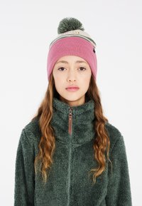Beanie a maglia rosa con una fascia a contrasto di colore crema e pompon verde. Il modello indossa una giacca in pile verde con colletto con zip.