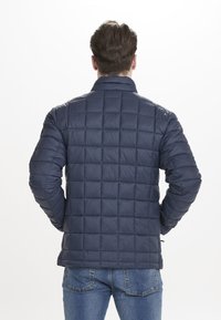 Marinblå puffjacka med kviltat mönster, har stående krage, ribbade ärmslut och dragkedja framtill, i kombination med blå jeans.