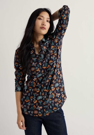 Woman with long black hair wearing a dark floral button-up shirt and dark jeans, posing with one arm raised behind her head.