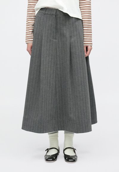 Jupe plissée grise en tissu texturé, arrivant au milieu du mollet. Associée à des chaussettes vert clair et des chaussures noires avec des accents décoratifs.