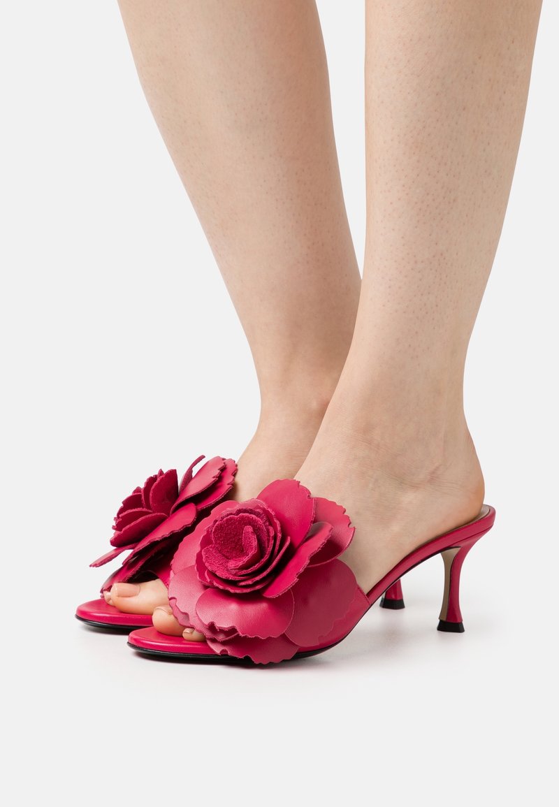 Pieds portant des sandales à talons ouvertes roses avec de grandes décorations florales sur le dessus sur fond blanc.
