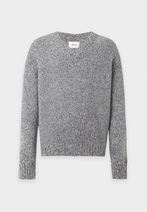 Homme portant un pull en maille gris à col en V, un jean droit bleu foncé avec une ceinture marron, et des mocassins marron foncé, debout devant un fond blanc.