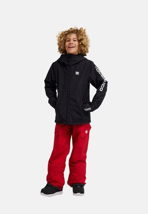 Kind met krullend haar dat een zwart capuchonjack en een rode broek draagt, staat met handen in de zakken glimlachend tegen een witte achtergrond.