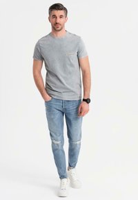 Camiseta de algodón gris con una pequeña impresión de "Original", combinada con jeans claros y desgastados y zapatillas blancas. Ajuste y estilo casual.