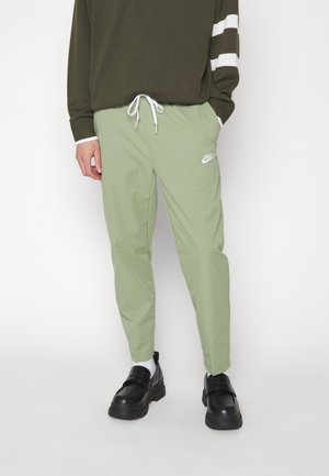 Model trägt hellgrüne Nike Jogginghosen mit weißem Kordelzug, dunkelgrünen Pullover mit weißen Streifen am Ärmel, weiße Socken und schwarze Schuhe.