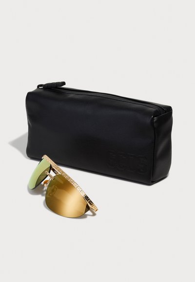 Sunglasses with green and gold-tinted lenses, gold patterned frame, alongside a black faux leather case embossed with "GCDS."