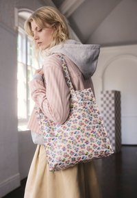 Blondínka v ružovej pruhovanej bunde a béžovej sukni nesie vnútri krémovú tašku na ramene s malými farebnými kvetinovými vzormi.