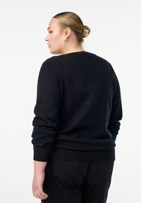 Schwarzer Strickpullover mit strukturiertem Wabenmuster, gerippten Bündchen und Saum, getragen mit schwarzen Hosen, von hinten gesehen.