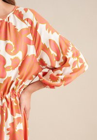 Langärmliges Kleid aus weißem Stoff mit kräftigem pink- und gelbfarbenem Blumenmuster. Elastischer Taillenbund und geraffte Details an den Ärmeln.