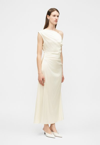 Γυναίκα που φοράει ένα μακρύ φόρεμα σε κρεμ απόχρωση, με έναν ώμο και ραμμένες λεπτομέρειες, στέκεται με τα χέρια χαλαρά στα πλάγια, φορώντας λευκά μυτερά τακούνια.