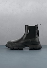 Bottine noire en cuir épais avec panneau latéral élastique, languette au talon et semelle épaisse de marque sur une surface grise.