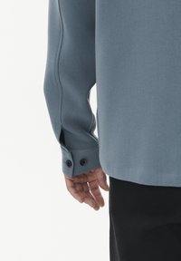 Långärmad, ljusblåskjorta med en slät textur. Har en knappslutning vid manschetterna och subtil sömdekoration längs ärmen. Svarta byxor synliga.