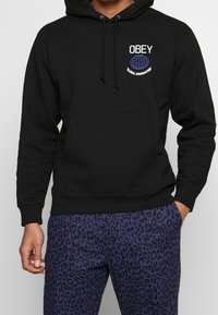 Černá mikina s kapucí, která má přední klokanní kapsu a nápis "OBEY GLOBAL DOMINATION", v kombinaci s fialovými kalhotami s leopardím potiskem.