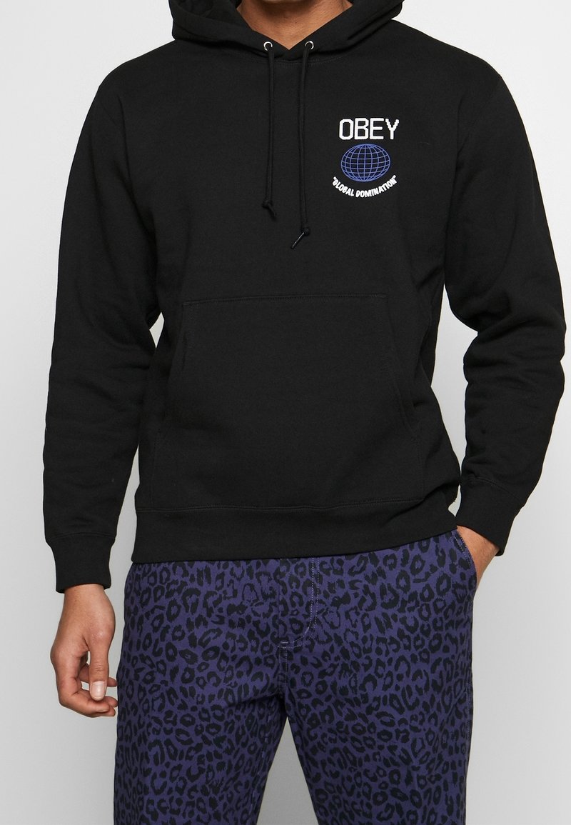 Černá mikina s kapucí, která má přední klokanní kapsu a nápis "OBEY GLOBAL DOMINATION", v kombinaci s fialovými kalhotami s leopardím potiskem.