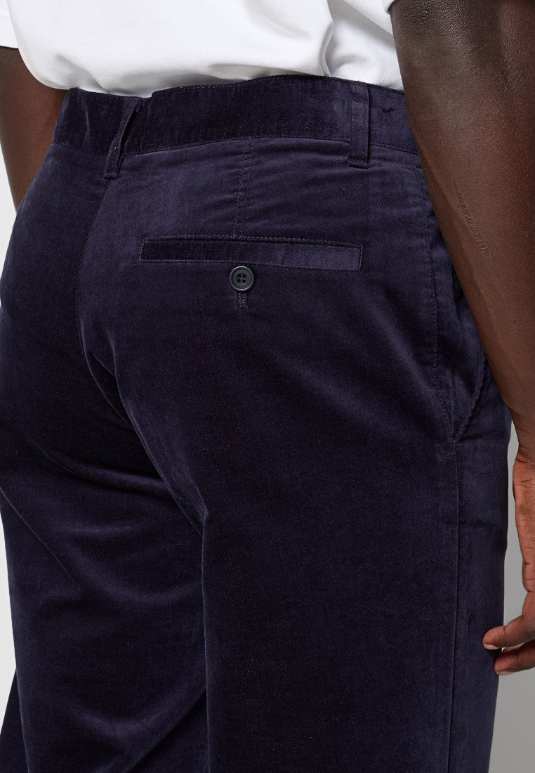 Close-up van donkerblauwe fluwelen broek met een achterzak met knopen, gedragen door een persoon in een wit overhemd, met focus op de heup en hand.