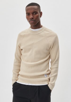 Pull en tricot côtelé beige avec col rond, tissu texturé, coupe slim, et étiquette brodée sur l'ourlet. Superposé sur une chemise blanche.